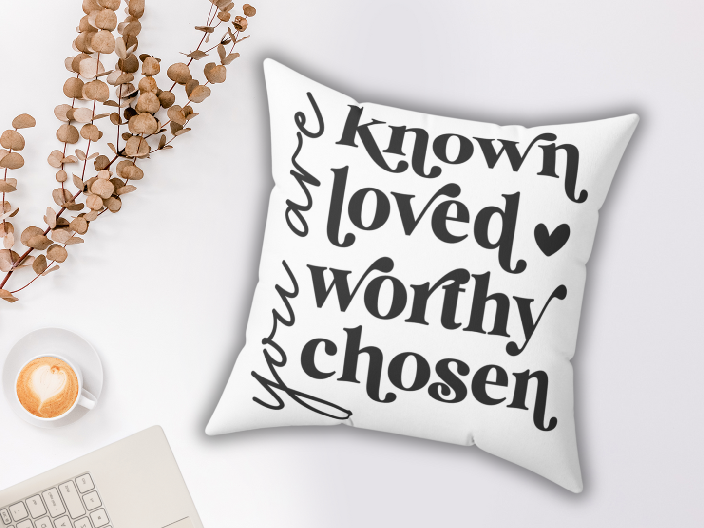 A pillow with the words "You are known, loved, worthy, chosen" in bold black script. Nearby are dried leaves, a cappuccino, and a laptop, creating a cozy and positive ambiance.
