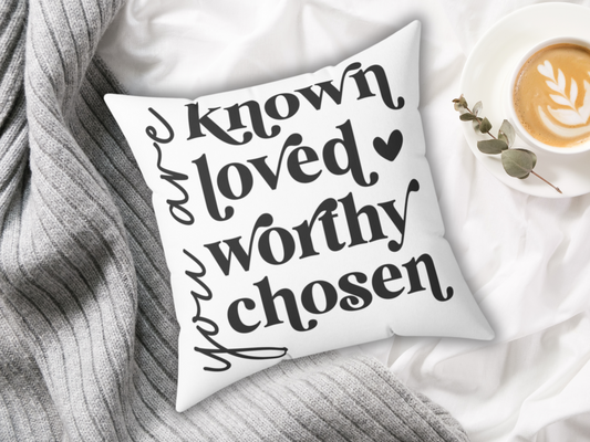 Decorative white pillow with the words "you are known loved worthy chosen" in bold black script. It's on a gray blanket next to a latte with latte art.