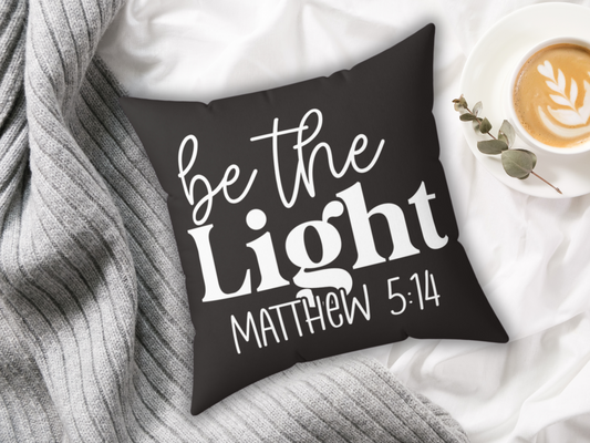 A black pillow with the phrase "be the Light, Matthew 5:14" in white text sits on a gray knit blanket next to a cappuccino with latte art on a white saucer.