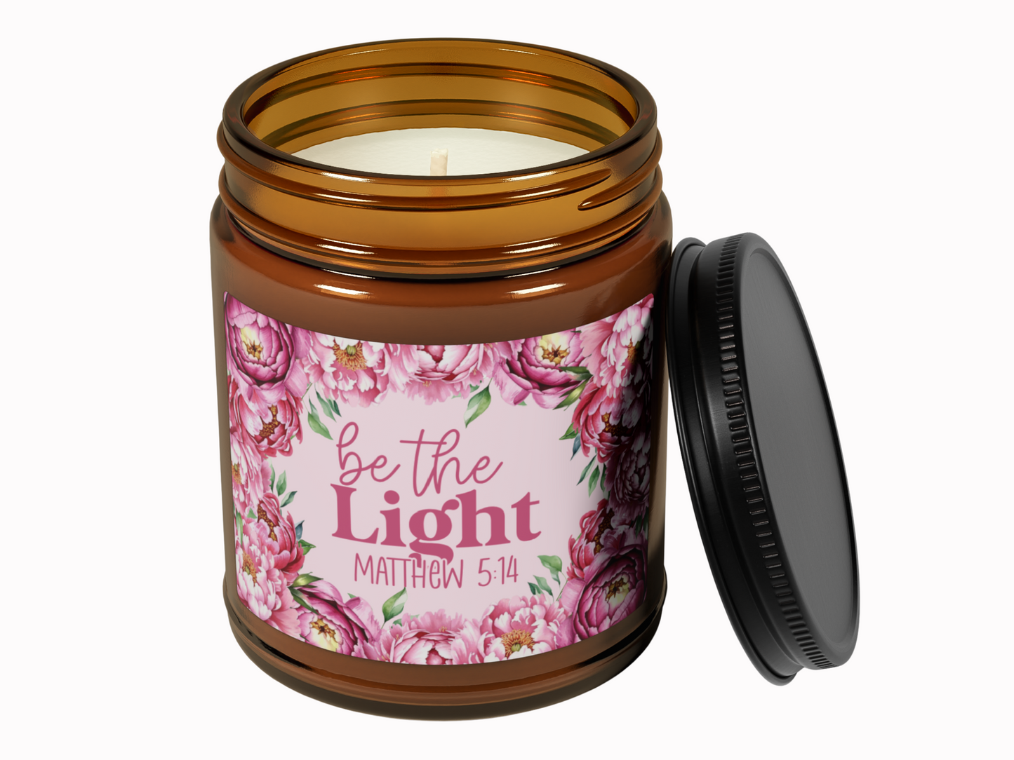 Brown glass candle jar with a black lid, featuring a floral label with pink flowers. Inscription reads “be the Light, Matthew 5:14,” conveying warmth.
