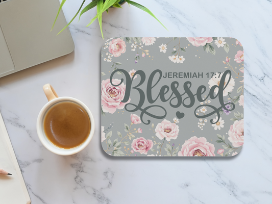 Mouse pad with "Blessed" and "Jeremiah 17:7" in script, surrounded by pink and white flowers. Nearby, a cup of coffee and a laptop on a marble surface convey a calming workspace setting.