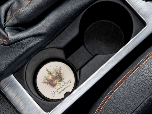 A car cup holder contains a coaster decorated with a watercolor crown and the text "Daughter of the King, 2 Corinthians 6:18." Leather detailing is visible.