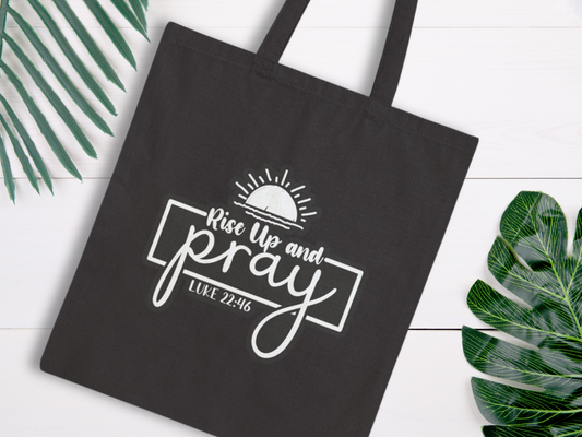 Black tote bag with "Rise Up and Pray, Luke 22:46" in white cursive and block letters. Sun icon above text. Tropical leaves on white wood background.