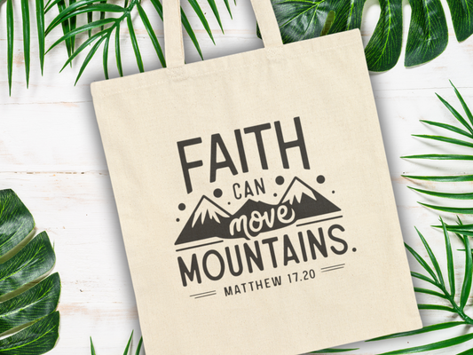 A beige tote bag on a white surface with green leaves. Printed in bold black text: "Faith can move mountains. Matthew 17:20" with mountain graphics.