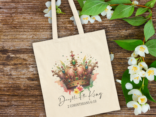Tote bag with a colorful crown and floral design, captioned "Daughter of the King, 2 Corinthians 6:18," lies on rustic wood with white flowers.