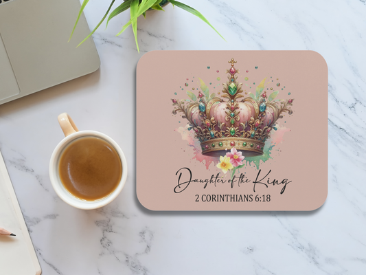 A pink mouse pad for laptop with a colorful jeweled crown and flowers. Text reads "Daughter of the King, 2 Corinthians 6:18." Nearby are a coffee cup, plant, and laptop.