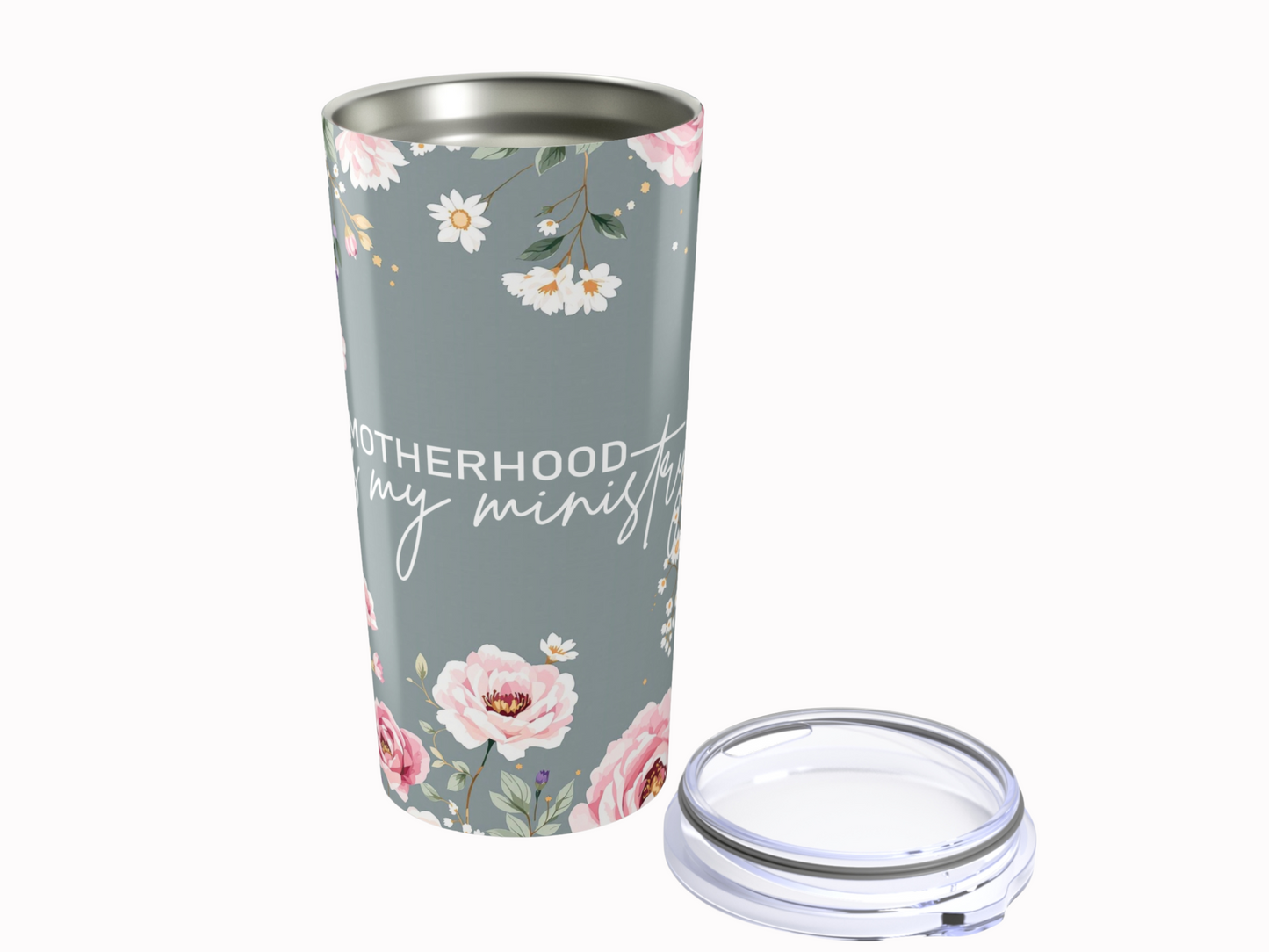 Gray tumbler with floral design and "Motherhood is my ministry" text. Features pink and white flowers. Lid is placed beside the tumbler. Elegant and cozy.