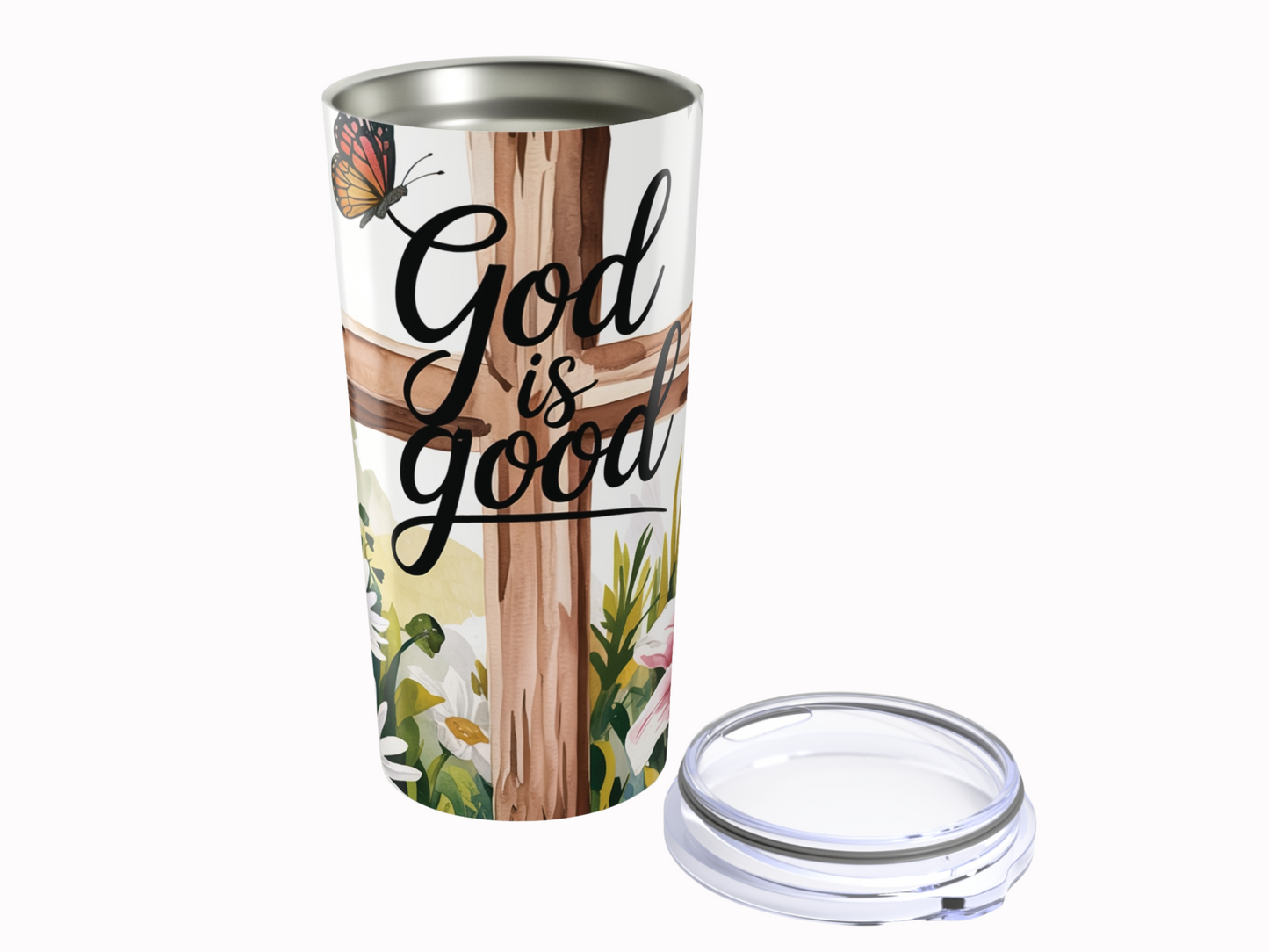 Tumbler with "God is good" text, featuring a wooden cross, a butterfly, and surrounding flowers. Lid placed beside it. Emotive and uplifting tone.