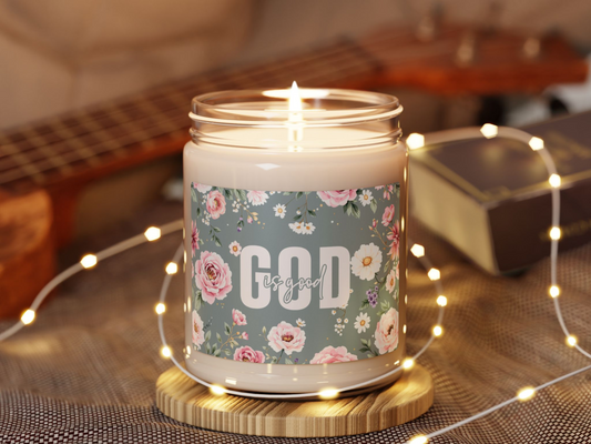 A lit candle labeled "God is good" with floral design sits on a wooden coaster. Nearby are twinkling fairy lights, a guitar, and a book, creating a warm, cozy ambiance.