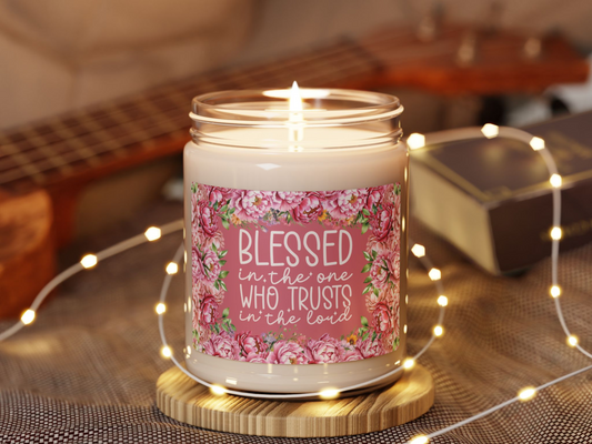 A lit candle labeled "Blessed in the one who trusts in the Lord" with floral design. Surrounded by soft fairy lights, a guitar, and a book, conveying warmth.