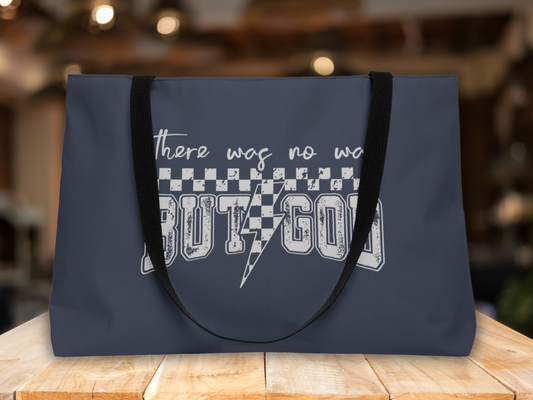 Navy blue tote bag with black straps on a wooden table, set against a blurred warm-toned indoor background, featuring “There was no way BUT GOD” text design.