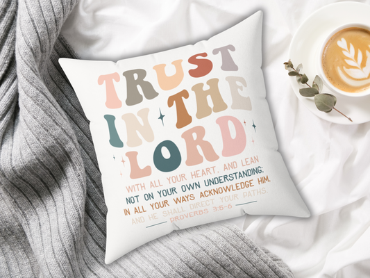 Christian scripture pillow with “Trust in the Lord” text, styled on white blanket with gray knit throw and latte in cup.
