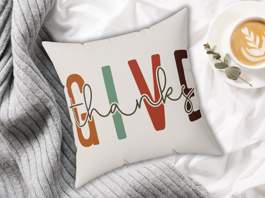 Cozy scene with a pillow reading "give thanks" in colorful letters on a gray blanket. Beside it, a cup of latte art coffee with a sprig of green leaves.