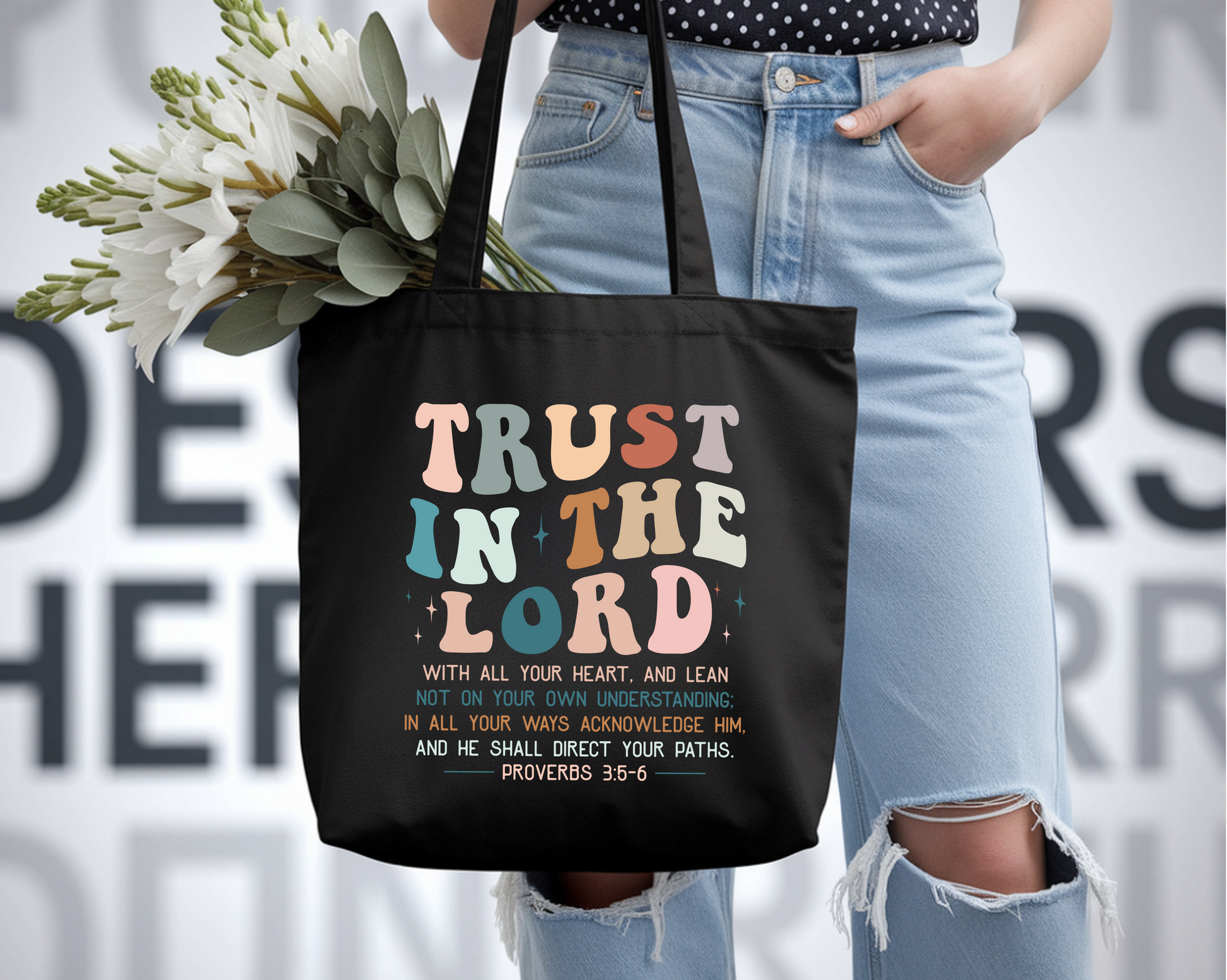 Person holding a black tote bag with colorful text and flowers, wearing light blue jeans.