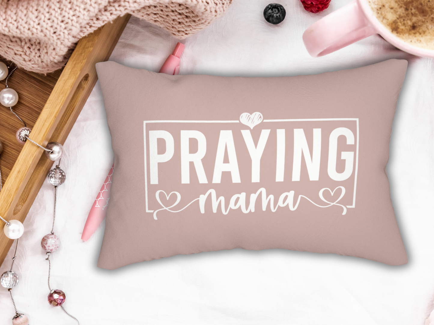 Blush pink lumbar pillow with white text “Praying Mama” in bold and script fonts with heart accents, styled with tray, coffee, and knit blanket.