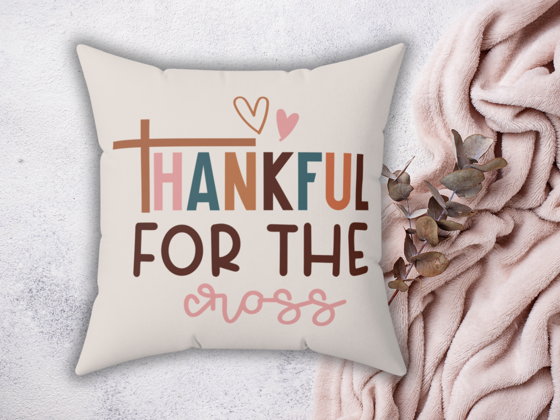 A square pillow with the text "Thankful for the cross" in colorful, playful fonts. The pillow rests on a pink blanket beside dried eucalyptus leaves.