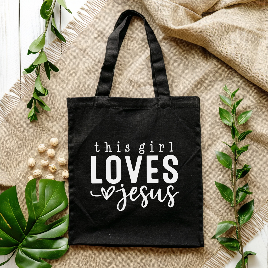 Black tote bag with 'this girl LOVES Jesus' text on a beige fabric background with green leaves.