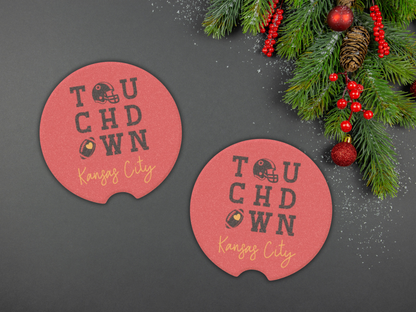 Two red coasters with 'Touchdown Kansas City' text on a dark surface with Christmas decorations.