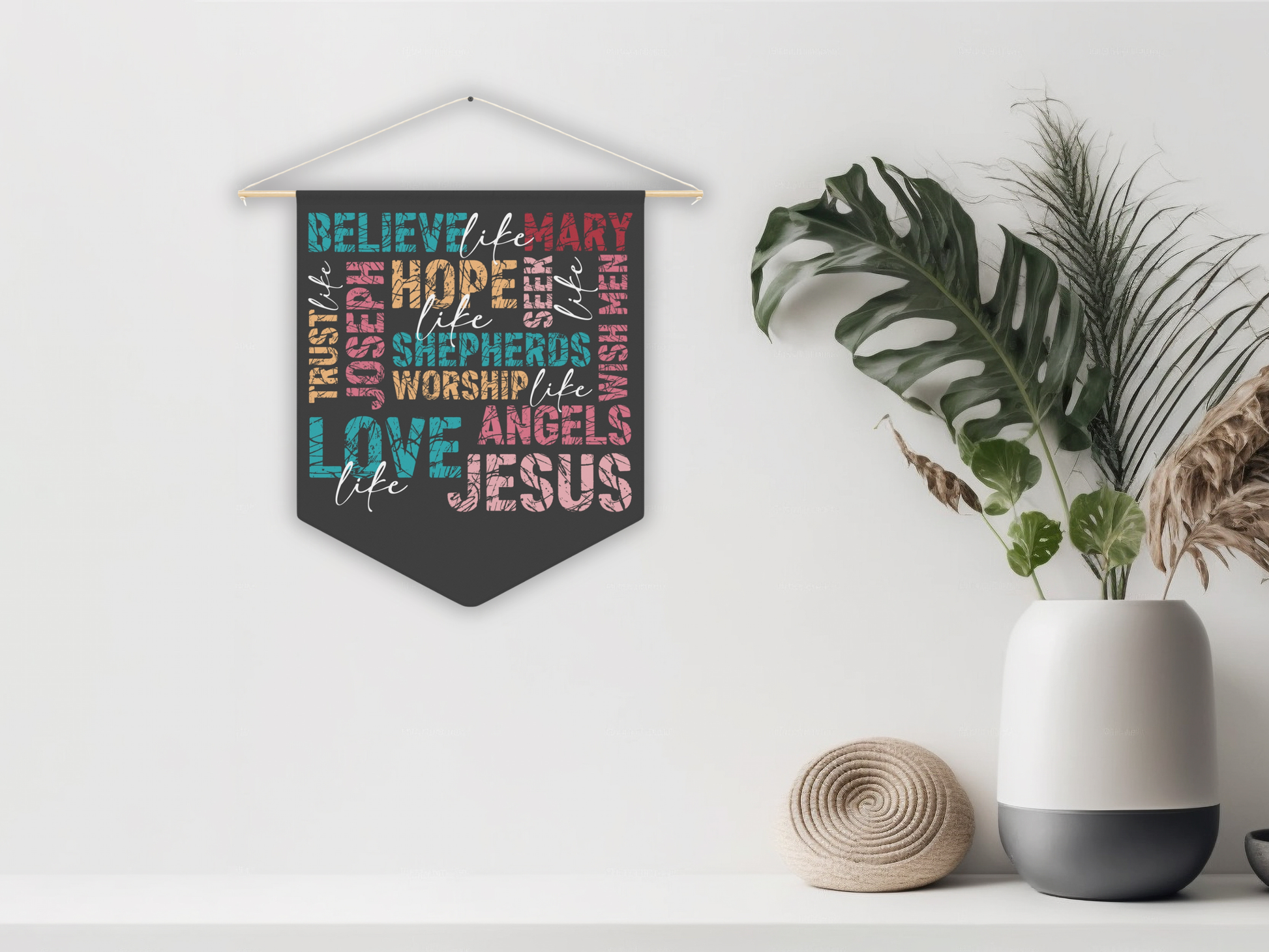 Decorative wall hanging with colorful text on a white wall with plants and a vase.