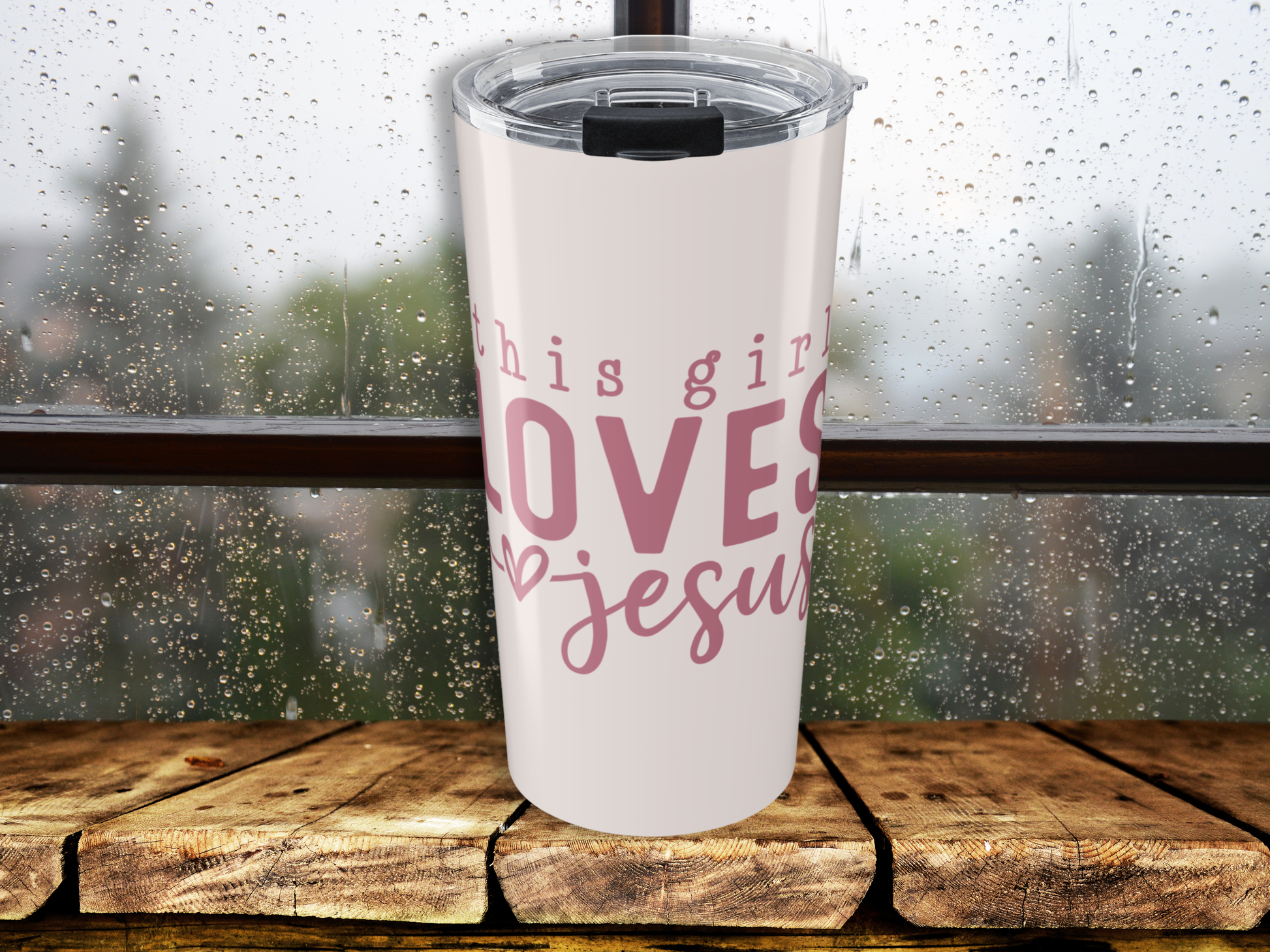 Tumbler with 'This girl loves Jesus' text on a wooden surface with a rainy window background