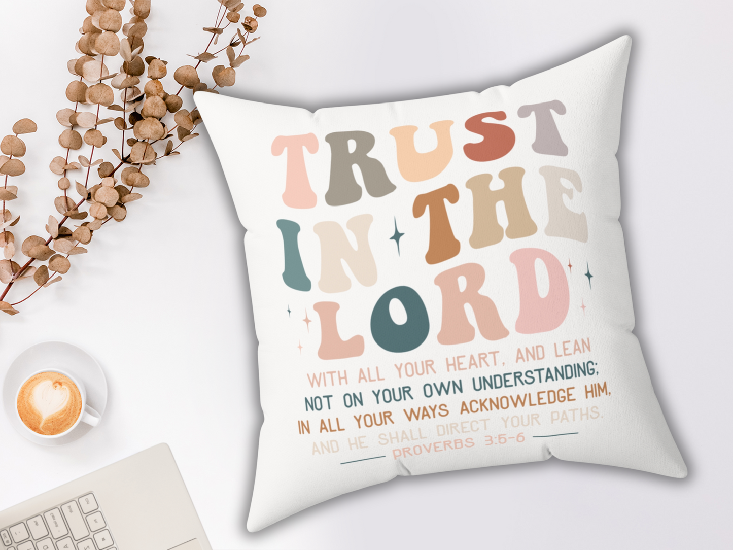 Inspirational Bible verse pillow with “Trust in the Lord” lettering, styled on white background with coffee, eucalyptus branches, and keyboard.