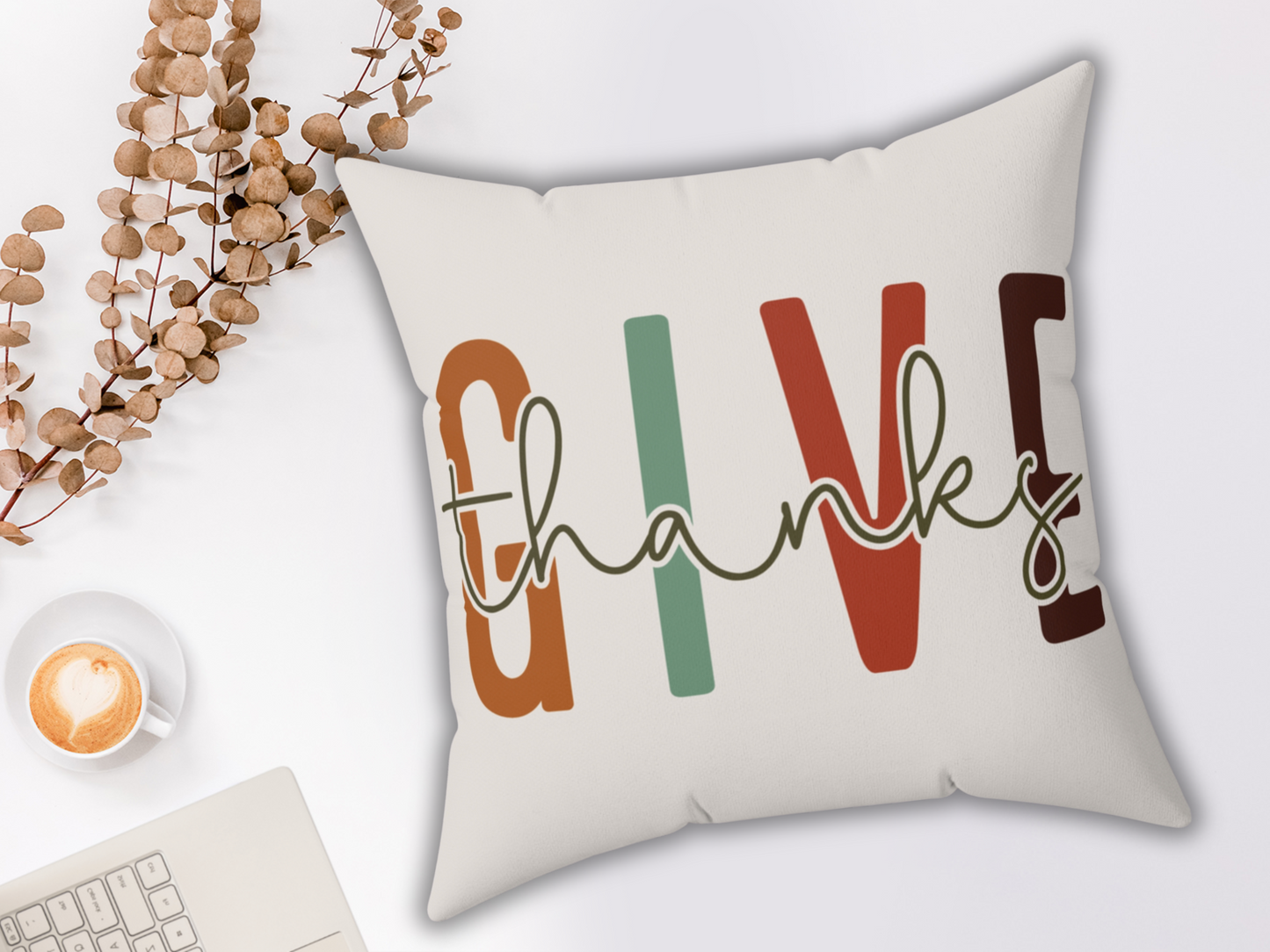 A decorative pillow features the words "Give Thanks" in colorful letters. Beside it are dried leaves, a cappuccino, and a laptop keyboard on a white surface.