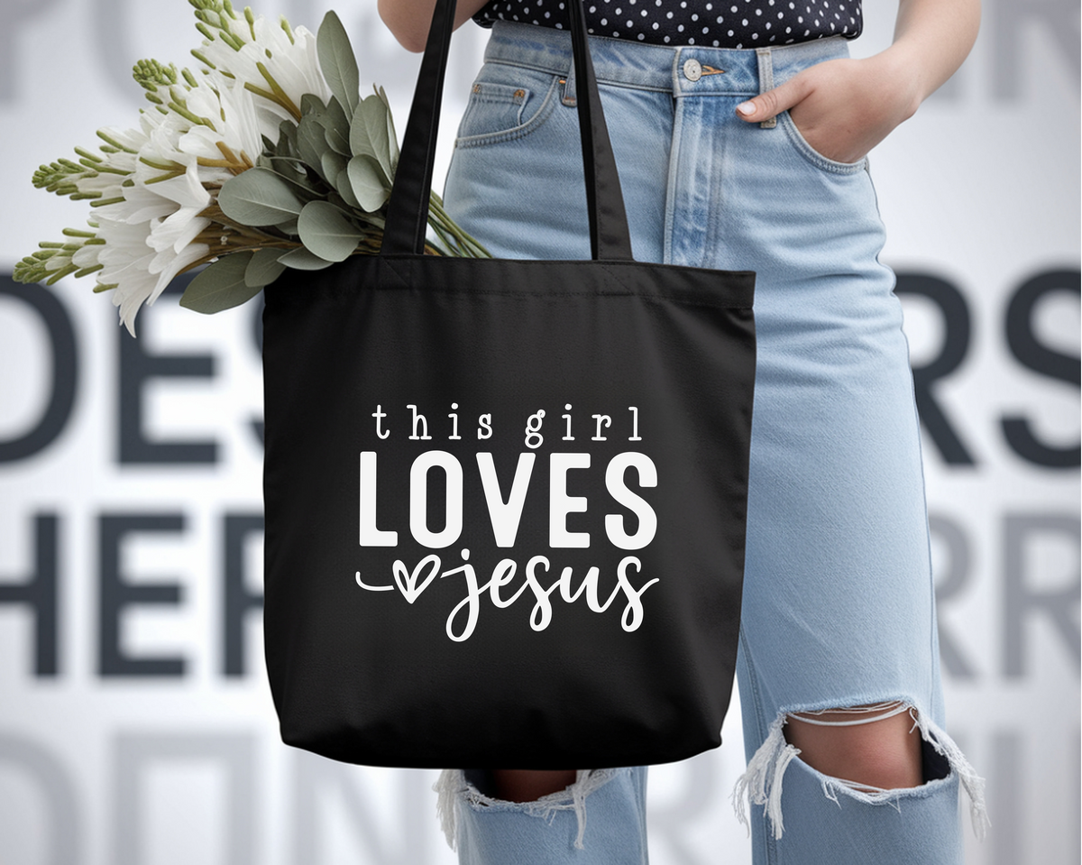 Person holding a black tote bag with 'this girl LOVES jesus' text, wearing light blue jeans.