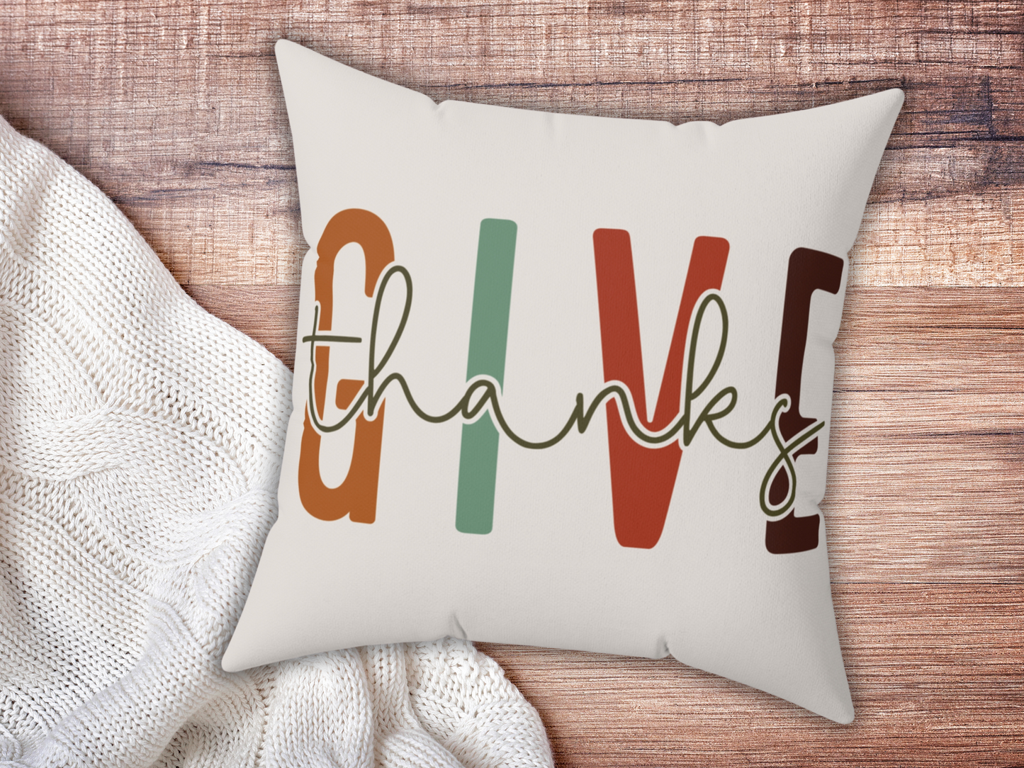 Cushion with "GIVE thanks" text in bold, colorful letters on a wooden surface, next to a cozy white knitted blanket. Warm, inviting feel.