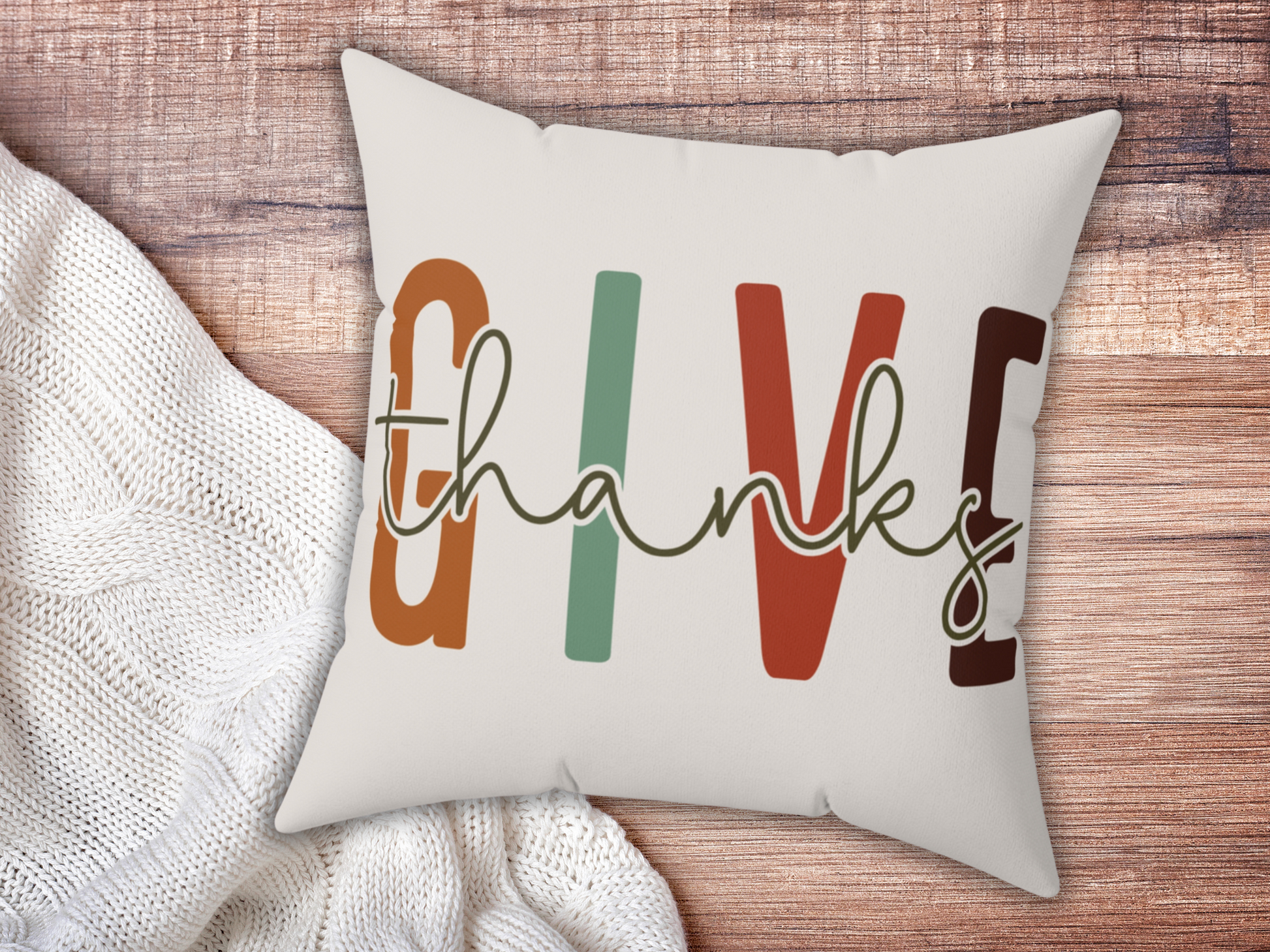 Cushion with "GIVE thanks" text in bold, colorful letters on a wooden surface, next to a cozy white knitted blanket. Warm, inviting feel.