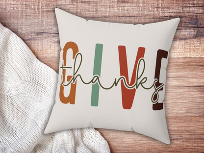 Cushion with "GIVE thanks" text in bold, colorful letters on a wooden surface, next to a cozy white knitted blanket. Warm, inviting feel.