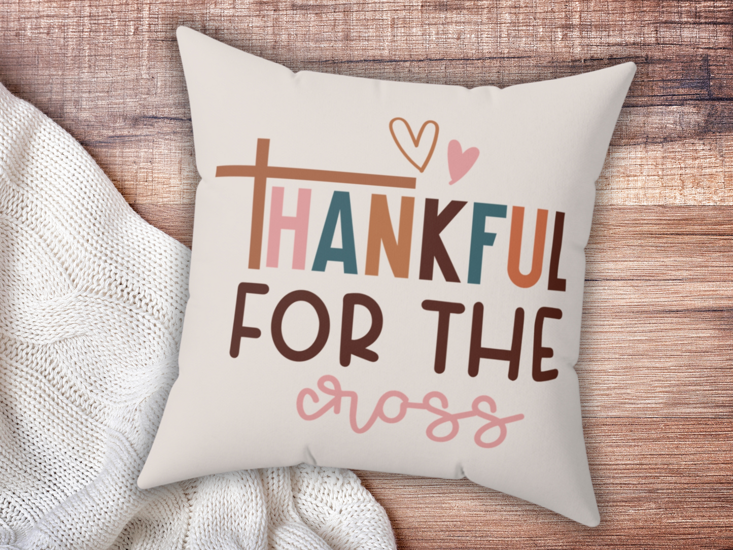 A decorative pillow on a wooden surface displays the text "Thankful for the Cross" in colorful, playful font with a heart design. Cozy and warm atmosphere.