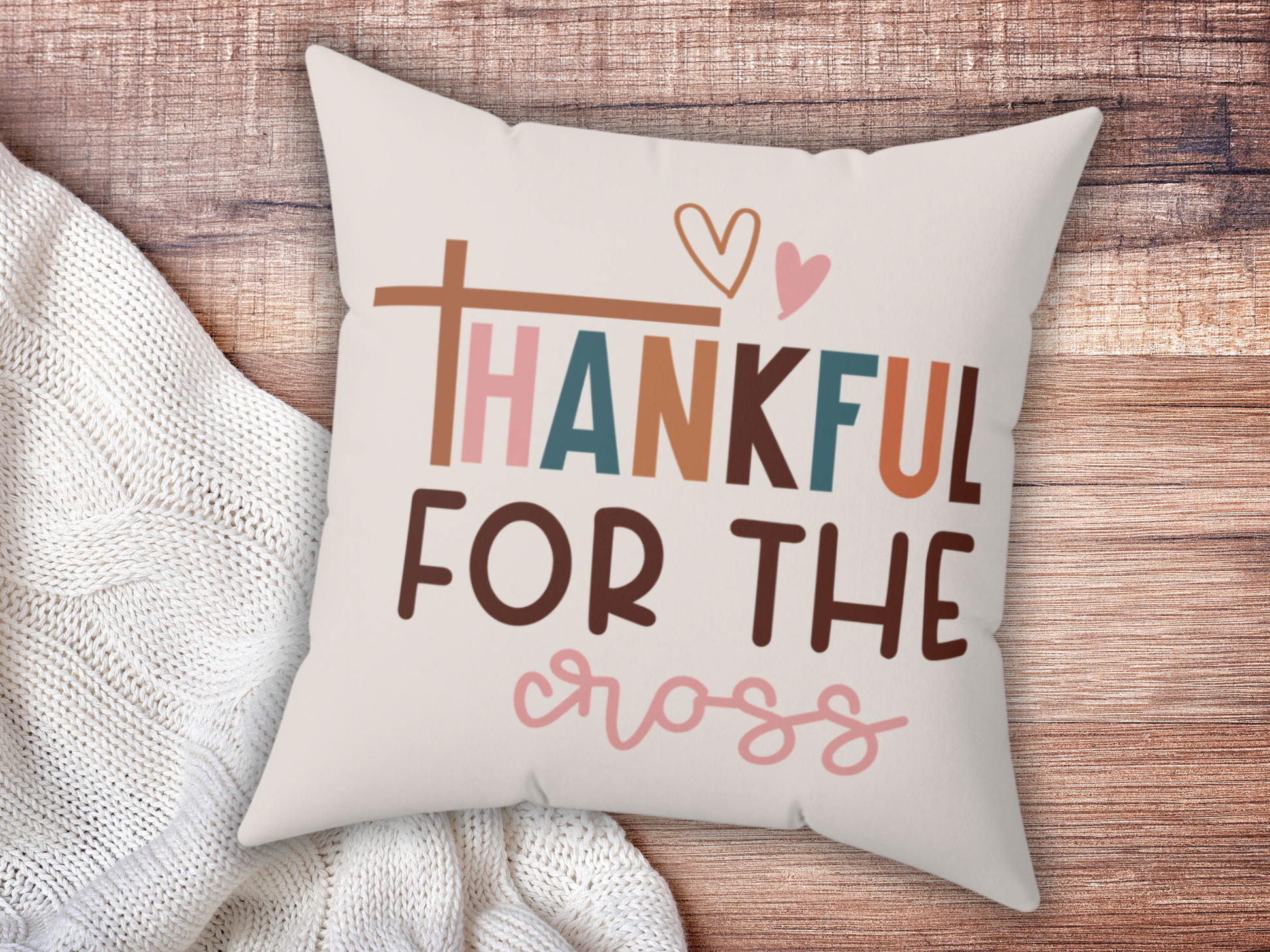 A decorative pillow on a wooden surface displays the text "Thankful for the Cross" in colorful, playful font with a heart design. Cozy and warm atmosphere.