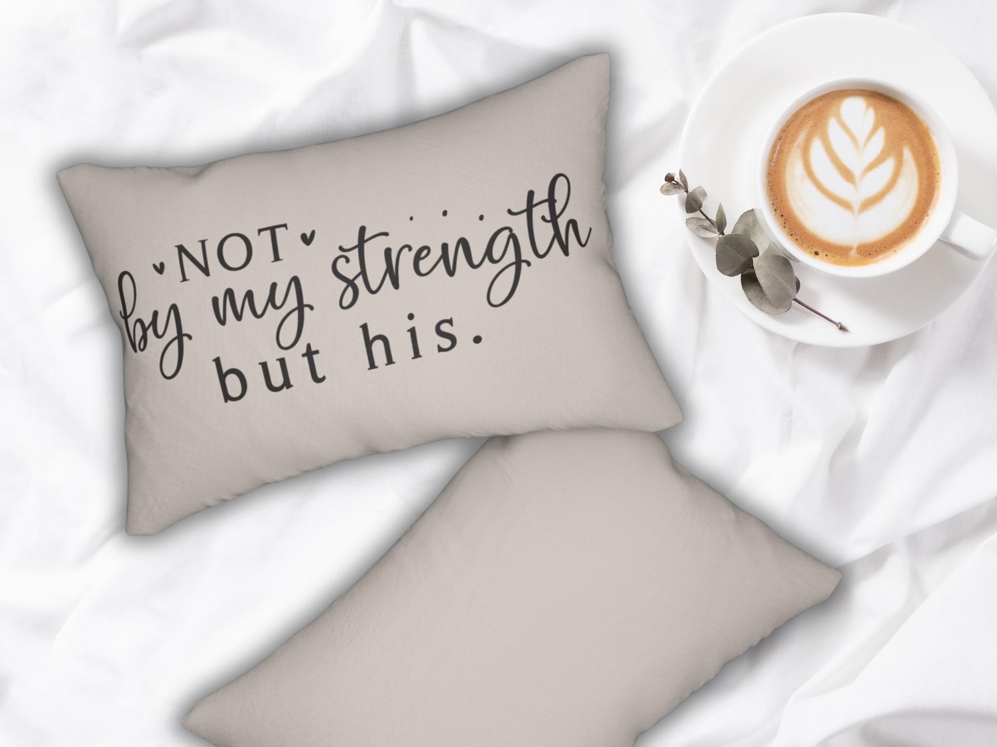 Cozy scene with a beige pillow displaying the phrase "Not by my strength but his" in black script. A cappuccino with latte art sits nearby on white fabric.