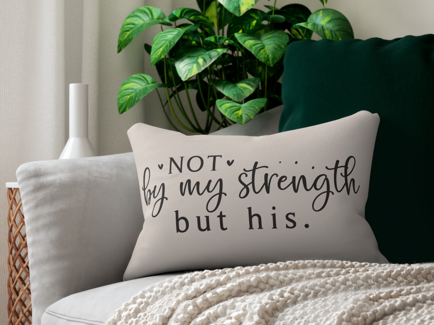 A cozy living room features a beige pillow with the phrase "Not by my strength but his." resting on a gray sofa. A green plant and textured blanket add warmth.