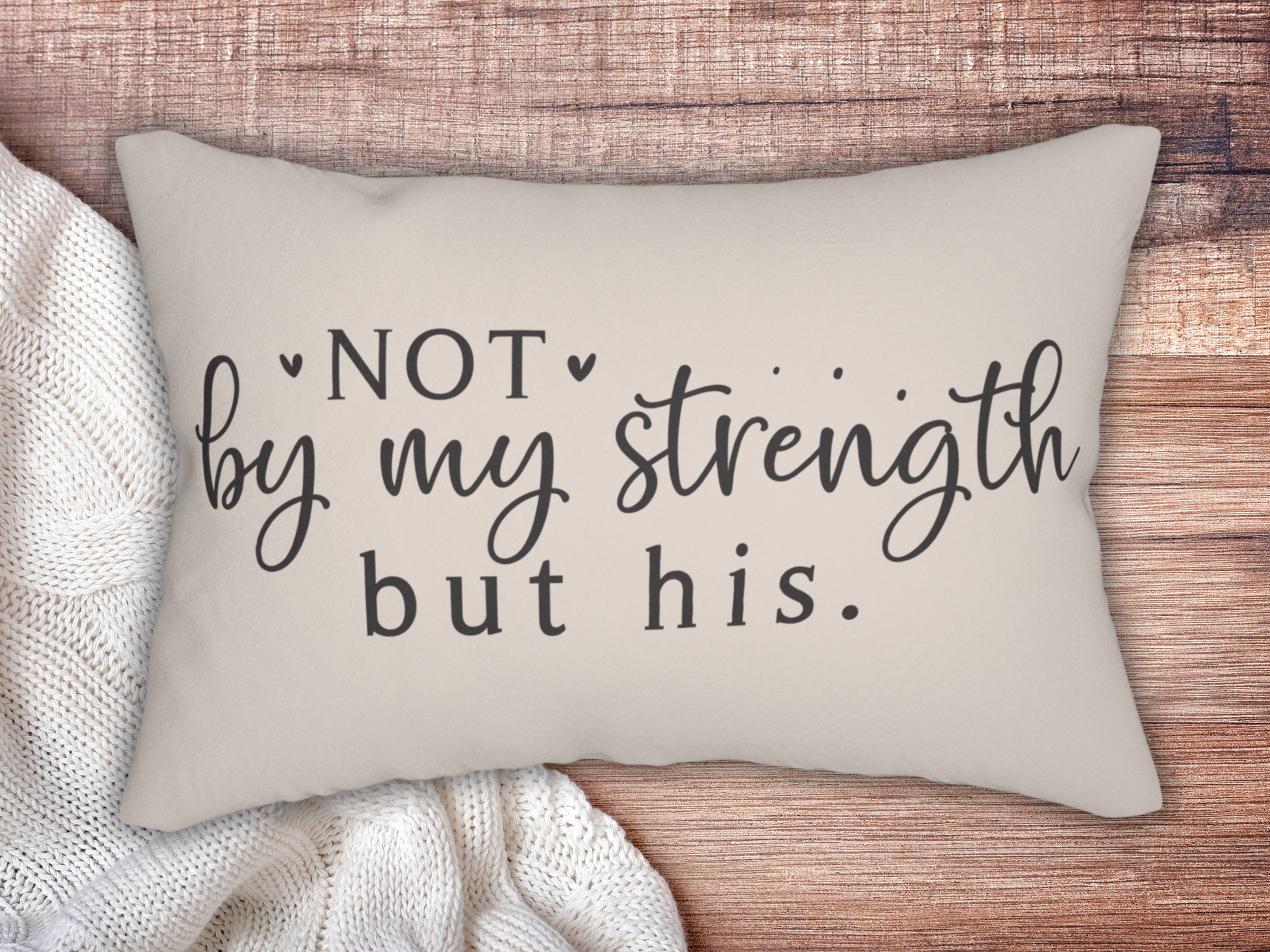 A beige pillow on a wooden surface shows a motivational quote: "Not by my strength but his." It rests beside a cozy white knit blanket, creating a warm and comforting tone.