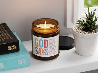 A lit candle in a brown jar on a windowsill, labeled "God says you are," alongside books and a small potted succulent plant. Cozy and thoughtful ambiance.