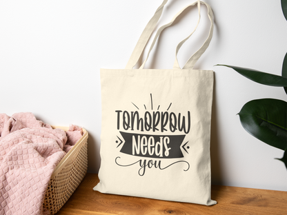 A cream tote bag with "Tomorrow Needs You" written in bold, playful font hangs on a wall. It rests near a wicker basket with a pink blanket, creating a cozy, inspirational vibe.