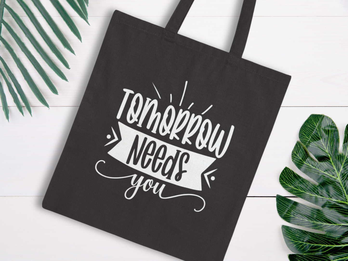 Black tote bag with white text "Tomorrow needs you" in playful script. Surrounded by green leaves on light wooden background, conveying positivity.