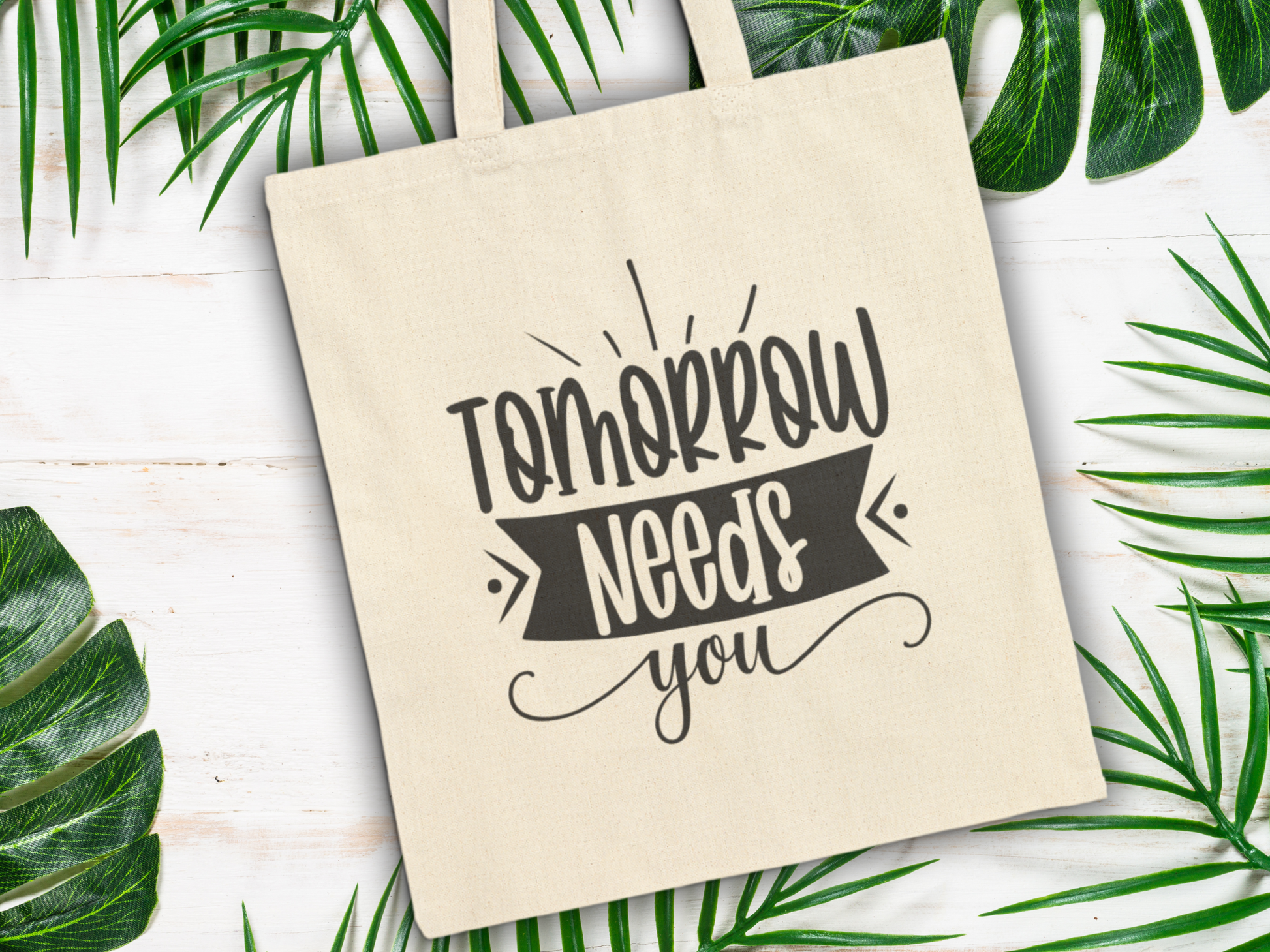 Beige tote bag with the text "Tomorrow Needs You" in bold black letters, surrounded by green tropical leaves on a white wooden surface.