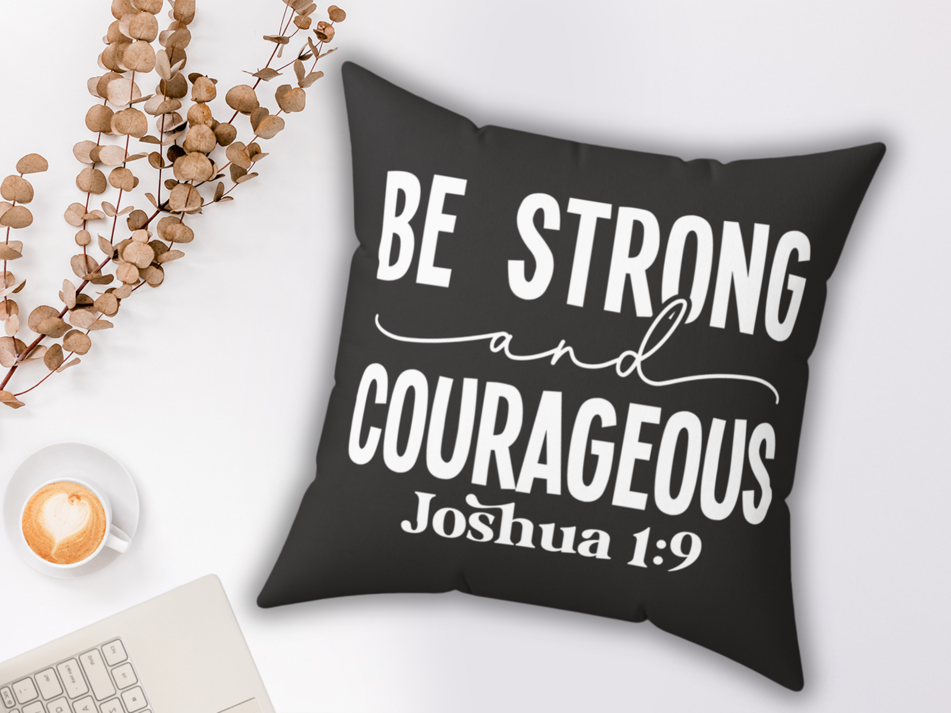 Black pillow with white text reads "Be Strong and Courageous, Joshua 1:9." Nearby are a latte with a heart design, eucalyptus sprigs, and a laptop, creating a cozy, inspirational setting.