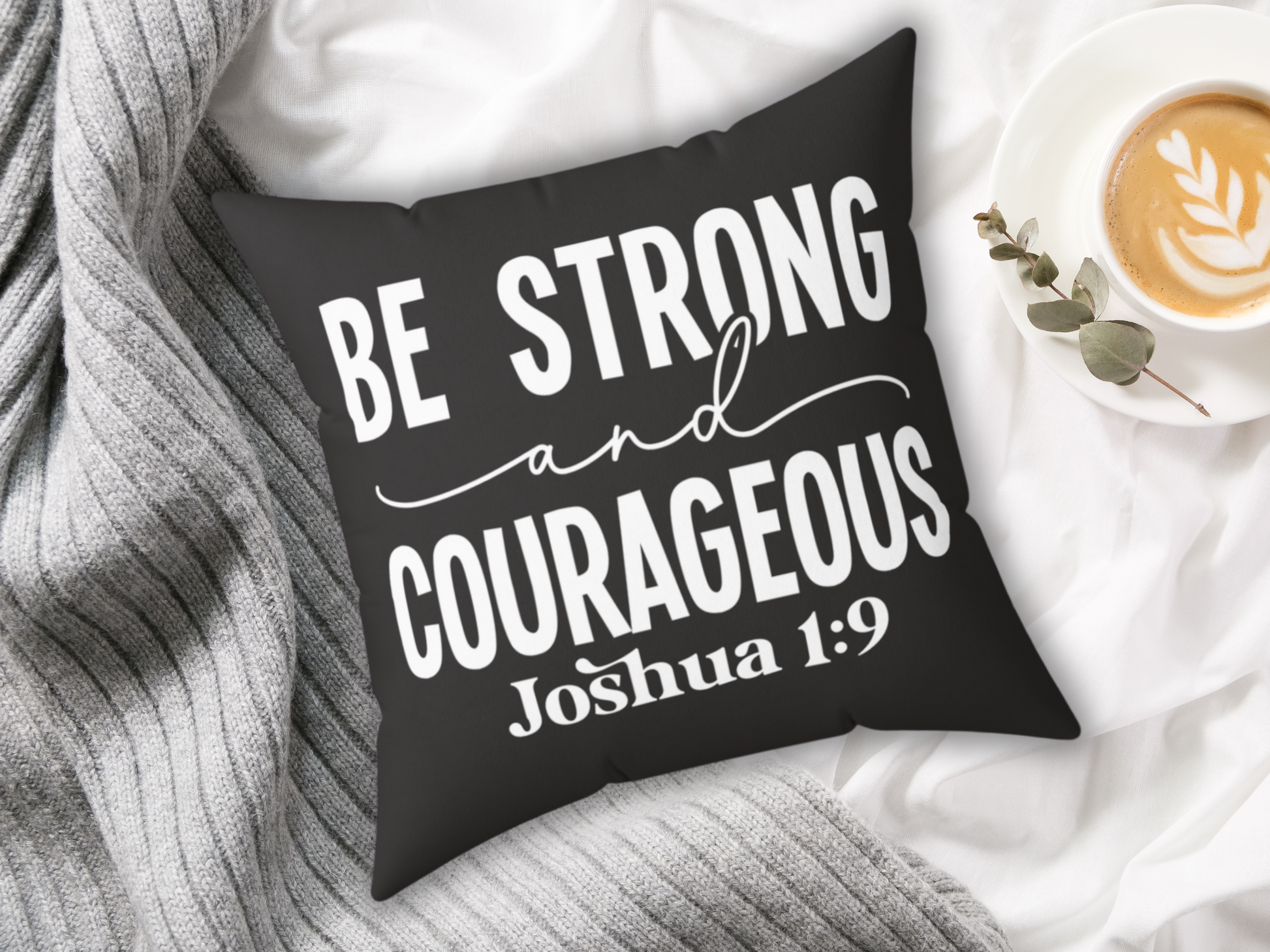 A black square pillow with white text reading "Be Strong and Courageous, Joshua 1:9" rests on a soft gray striped blanket. Nearby is a cappuccino in a white cup, adorned with latte art, and a sprig of eucalyptus, creating a cozy, uplifting atmosphere.