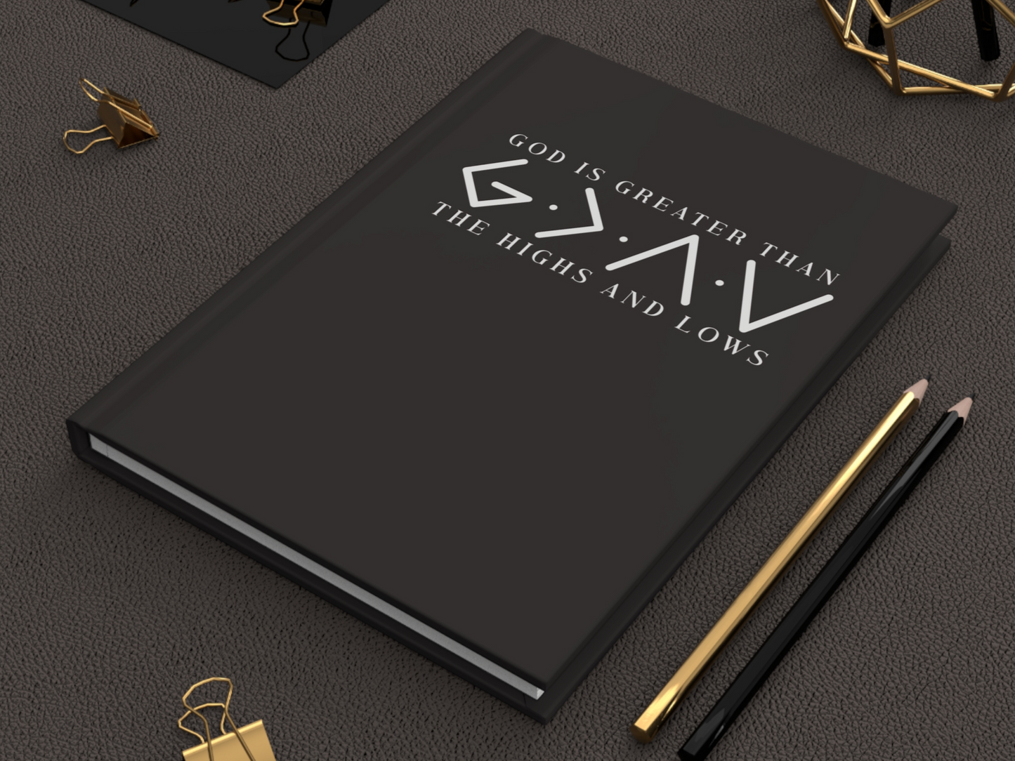 A black journal with the slogan "God is Greater Than the Highs and Lows" in white text on a textured surface. Gold and black pencils and clips are nearby.