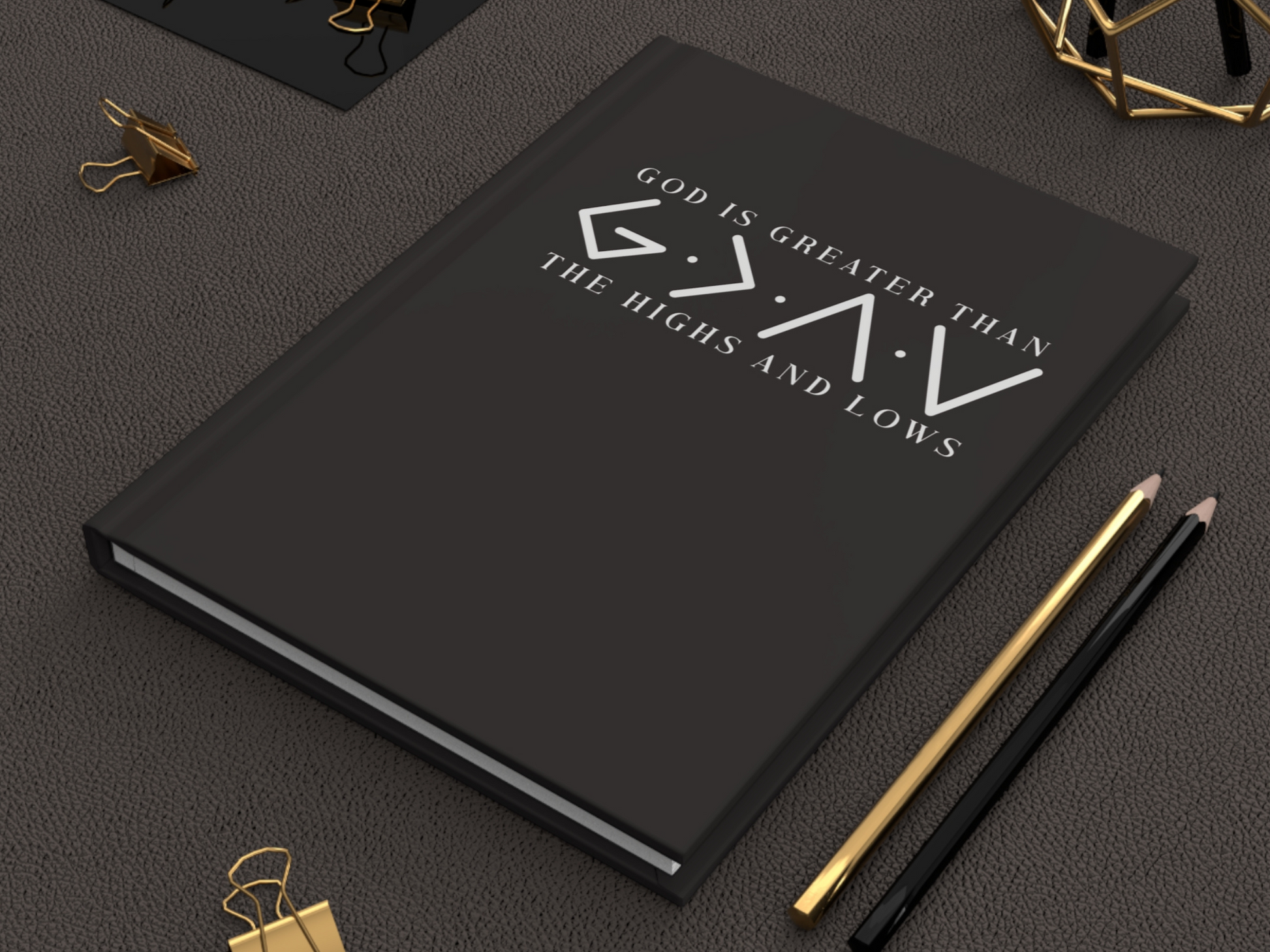 A black journal with the slogan "God is Greater Than the Highs and Lows" in white text on a textured surface. Gold and black pencils and clips are nearby.
