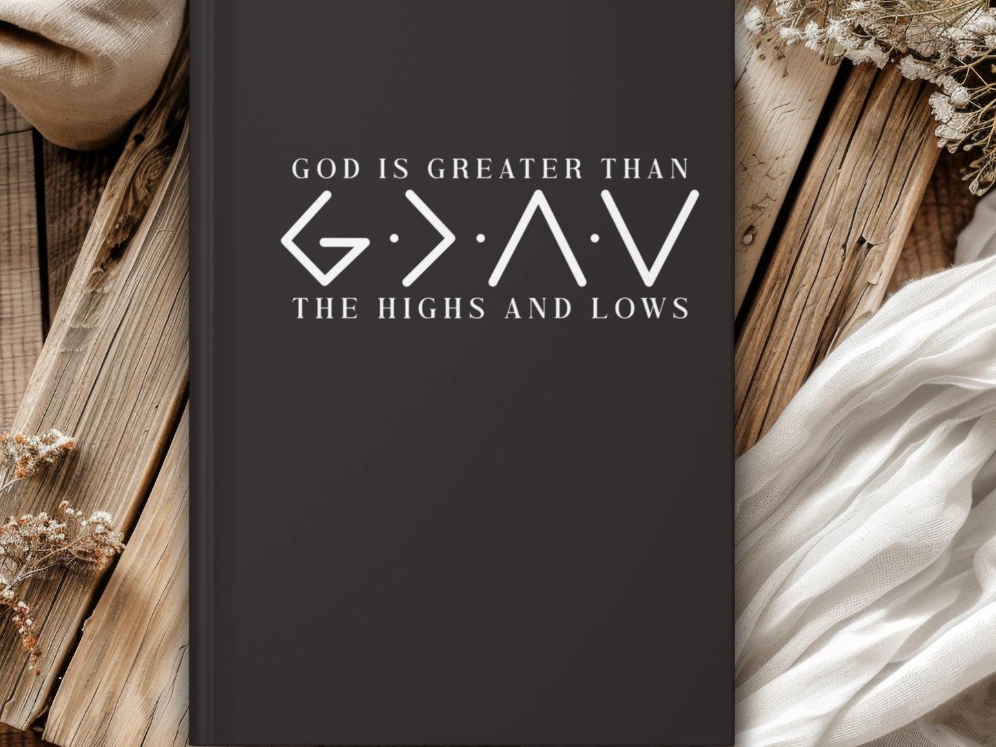Black journal cover with the text "God is greater than the highs and lows." Rustic wooden background with white fabric and dried flowers.