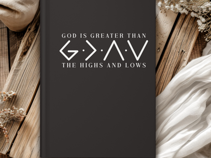 Black journal cover with the text "God is greater than the highs and lows." Rustic wooden background with white fabric and dried flowers.