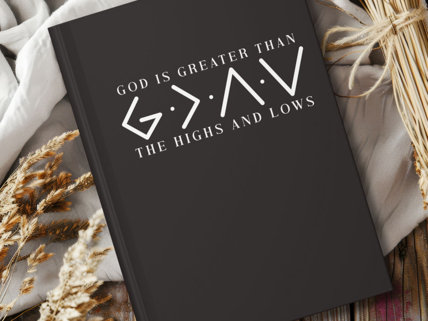 A black book cover reads "God is Greater Than the Highs and Lows" with symbols, lying on a white cloth. Surrounding it are wheat stalks and wooden planks.