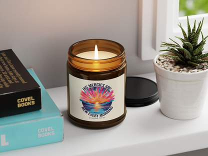 Lit candle in amber jar with "His Mercies Are New Every Morning" label beside books and a small potted succulent on a windowsill. Calm, cozy atmosphere.