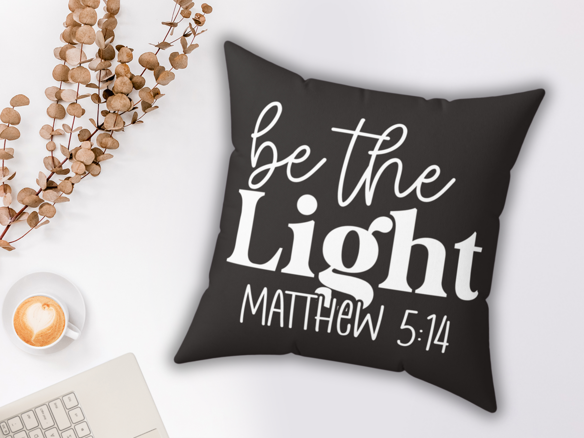 A black pillow with white cursive text reads "be the Light Matthew 5:14." Nearby are a latte with a heart design, dried branches, and a laptop corner. Cozy and inspirational.