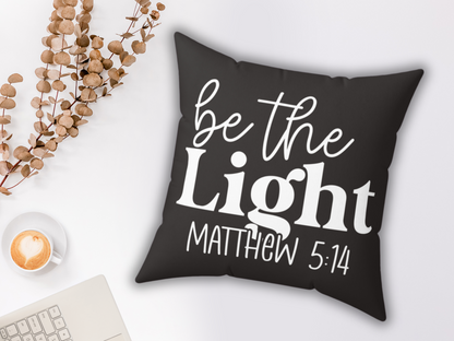 A black pillow with white cursive text reads "be the Light Matthew 5:14." Nearby are a latte with a heart design, dried branches, and a laptop corner. Cozy and inspirational.