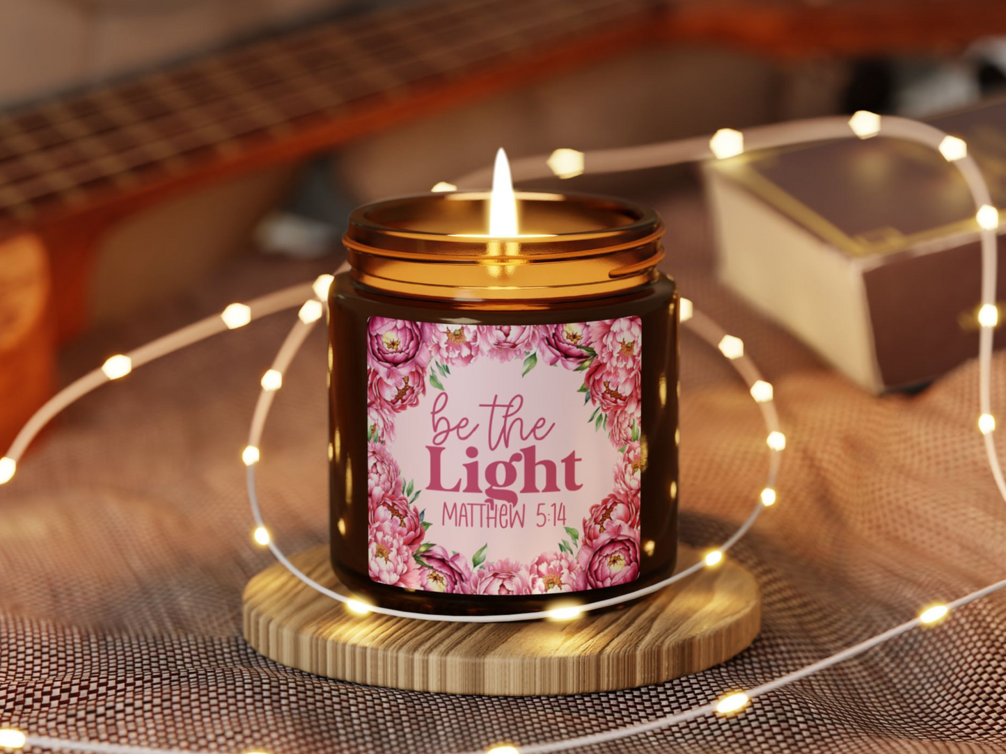 A lit candle on a wooden coaster, labeled with floral designs and the phrase "be the light Matthew 5:14." Surrounded by soft, glowing fairy lights.